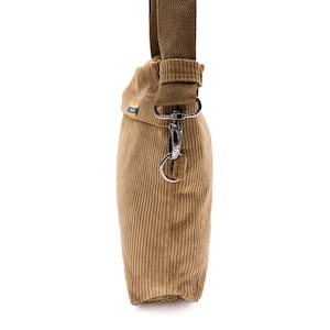 Op de afbeelding: Een bruin corduroy crossbody tas met een bruine canvas riem. De tas heeft een rechthoekige vorm met verticale corduroy ribbels. Een zilverkleurige metalen sluiting verbindt de riem met de tas.