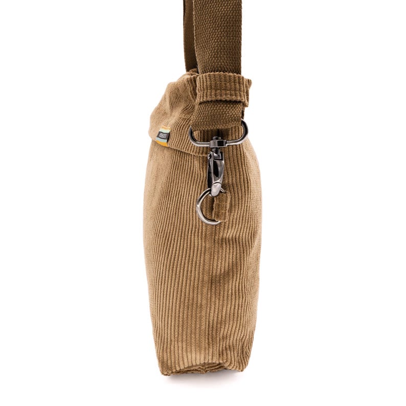 Op de afbeelding: Een bruin corduroy crossbody tas met een bruine canvas riem. De tas heeft een rechthoekige vorm met verticale corduroy ribbels. Een zilverkleurige metalen sluiting verbindt de riem met de tas.