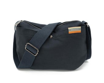 Op de afbeelding: Een marineblauwe messenger bag met een lange verstelbare riem en een zilveren gesp. De tas heeft een ritssluiting aan de bovenkant en een klein label met het woord "seasara" in witte tekst. De tas is gemaakt van een duurzame stof.