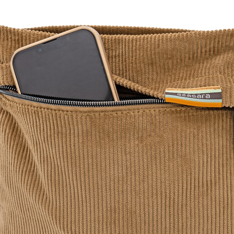 Op de afbeelding: Close-up van een bruin corduroy tas met een zwarte rits en een smartphone in de zak. De tas heeft een label met het woord "seasara" in witte tekst op een gestreepte achtergrond van oranje, blauw en groen.