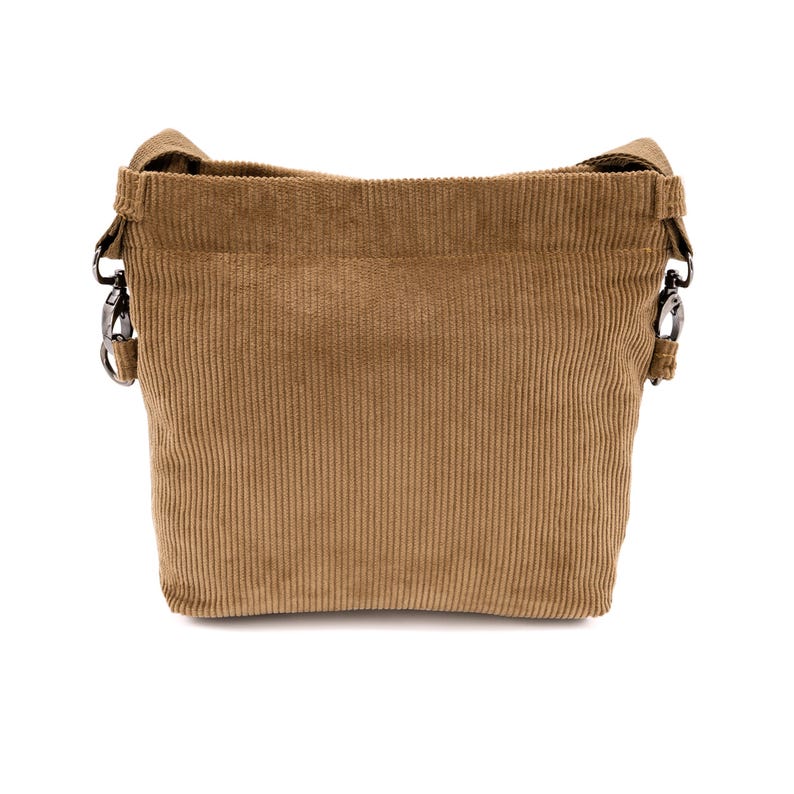 Op de afbeelding: Een bruine corduroy emmer tas met een schouderriem. De tas heeft een rechthoekige vorm met verticale corduroy ribbels. De riem is met metalen sluitingen aan de tas bevestigd. De tas is gemaakt van een zachte, getextureerde stof.