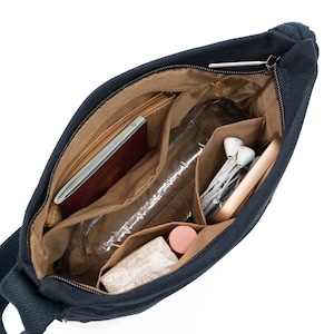 May include: A dark blue canvas shoulder bag with multiple pockets and compartments. The bag is open and filled with various items, including a passport, a water bottle, headphones, and a phone.