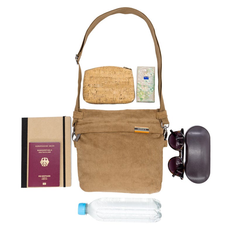 Op de afbeelding: Een bruine corduroy crossbody tas met bijpassende riem, een kurken etui, zonnebril, een notitieboek, een paspoort, een klein pakje tissues en een waterfles. De tas heeft een klein oranje en wit label.