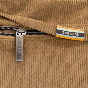 Op de afbeelding: Close-up van een bruin corduroy stof met een zwarte rits en een metalen ritstrekker. De ritstrekker en een stoffen label tonen beide de merknaam "seasara". Het label heeft strepen in teal, oranje en wit.