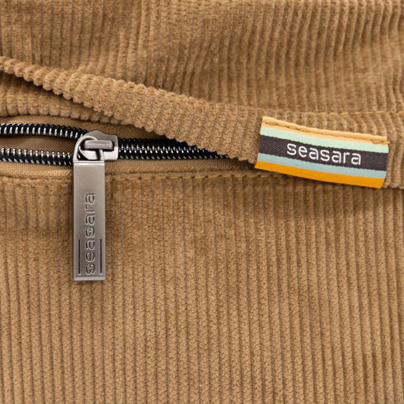Op de afbeelding: Close-up van een bruin corduroy stof met een zwarte rits en een metalen ritstrekker. De ritstrekker en een stoffen label tonen beide de merknaam "seasara". Het label heeft strepen in teal, oranje en wit.