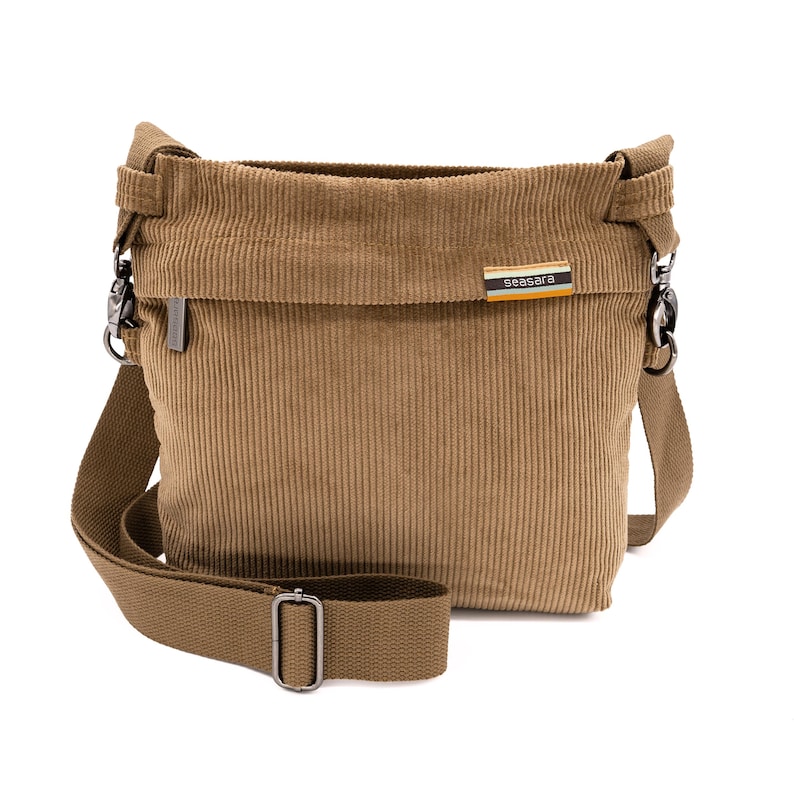 Op de afbeelding: Een bruin corduroy crossbodytasje met een lange verstelbare riem en zilverkleurige hardware. De tas heeft een rechthoekige vorm met een overslag en een klein label met de tekst "seasara".