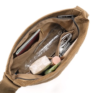 Op de afbeelding: Opengeklapte, bruin-beige corduroy schoudertas met inhoud. Binnenin zitten een paspoort, waterfles, oortelefoons en andere persoonlijke spullen. De tas heeft een ritssluiting en meerdere binnenvakken.