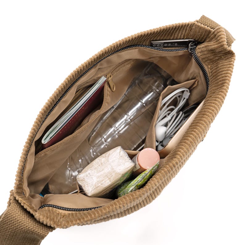 Op de afbeelding: Opengeklapte, bruin-beige corduroy schoudertas met inhoud. Binnenin zitten een paspoort, waterfles, oortelefoons en andere persoonlijke spullen. De tas heeft een ritssluiting en meerdere binnenvakken.