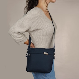 May include: A dark blue crossbody bag with silver hardware and a small, rectangular label on the front. The bag is being worn by a person wearing a light beige sweater and blue jeans.