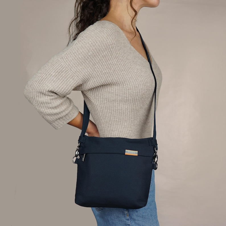 May include: A dark blue crossbody bag with silver hardware and a small, rectangular label on the front. The bag is being worn by a person wearing a light beige sweater and blue jeans.