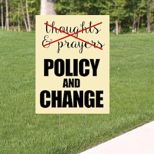 May include: A beige yard sign with black text that reads "thoughts & prayers" with a red line through it. Below the text is "POLICY AND CHANGE" in black.