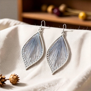 Puede incluir: Un par de pendientes plateados con forma de hoja. Los pendientes presentan un diseño delicado con un marco plateado y detalles azules en forma de hilo. El diseño es simétrico, con una serie de pequeños círculos a lo largo del borde exterior.