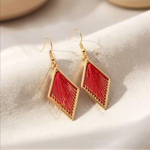 Puede incluir: Pendientes dorados en forma de diamante con un diseño de hilo rojo. Los pendientes tienen un patrón geométrico y un cierre de gancho. Los pendientes se muestran sobre una tela blanca.