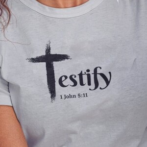 May include: A light gray t-shirt with the word "Testify" and a cross graphic. The text "1 John 5:11" is below the word "Testify". The person wearing the shirt has curly auburn hair and is smiling.