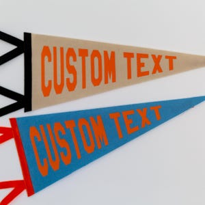 Könnte beinhalten: Zwei dreieckige Filzwimpel mit orangefarbenem Text CUSTOM TEXT. Der obere Wimpel ist beige mit schwarzen Bändern, der untere Wimpel ist blau mit roten Bändern. Beide Wimpel haben eine spitze Form und eine deutliche Beschriftung auf der Vorderseite.
