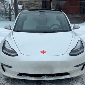 May include: A white electric car with a black roof and a red maple leaf emblem on the front. The car has sleek headlights and is parked in a snowy environment.