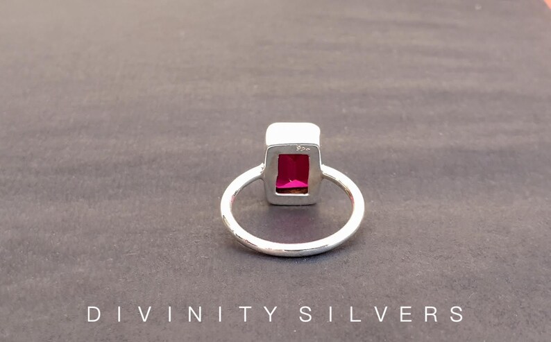 Peut inclure: Une bague en argent avec une pierre pr&eacute;cieuse rectangulaire rose fonc&eacute; sertie dans une b&eacute;li&egrave;re en argent. La bague est sur une surface grise.