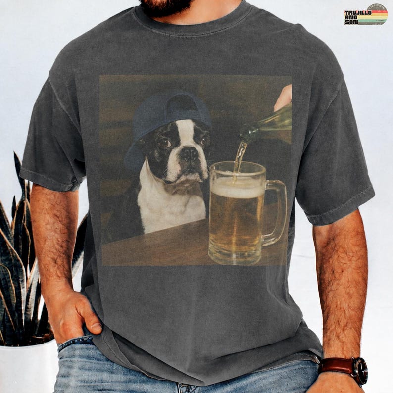 May include: A charcoal gray t-shirt featuring a graphic print of a Boston Terrier wearing a dark blue baseball cap, watching as a hand pours a golden beer into a glass mug. The shirt has a vintage-style washed finish and short sleeves.