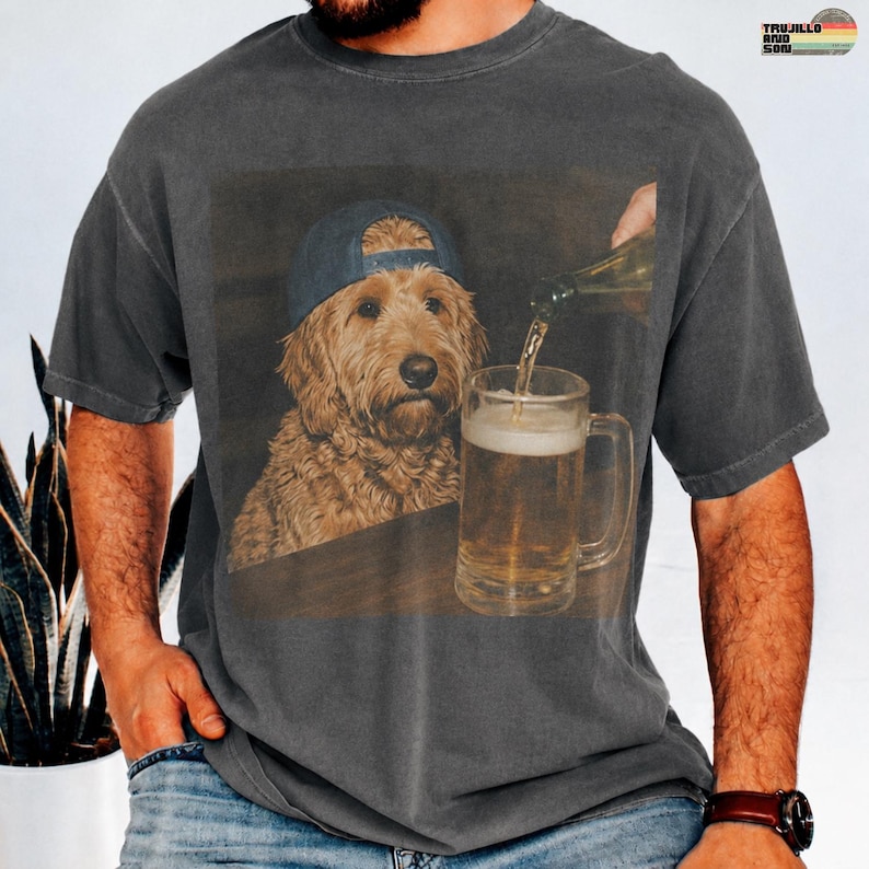 May include: A charcoal gray short-sleeve t-shirt featuring a graphic print of a golden-colored dog wearing a blue baseball cap. The dog is positioned next to a glass beer mug being filled with a golden liquid from a bottle held above the glass.