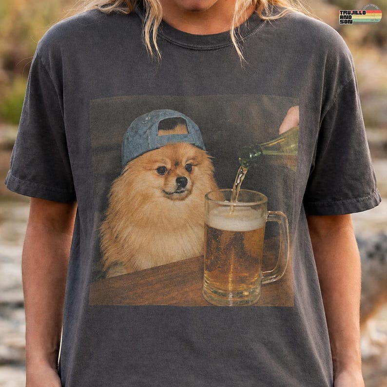 May include: A charcoal gray t-shirt featuring a graphic print of a fluffy tan Pomeranian dog wearing a blue baseball cap. The dog is positioned next to a glass beer mug being filled from a bottle. The shirt has a crew neckline and short sleeves.