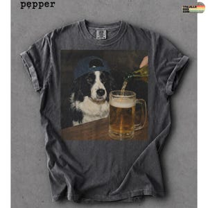 May include: A charcoal gray short-sleeve t-shirt featuring a graphic print of a black and white dog wearing a blue baseball cap, watching as liquid is poured from a bottle into a glass beer mug. The word pepper is printed in the top left corner.