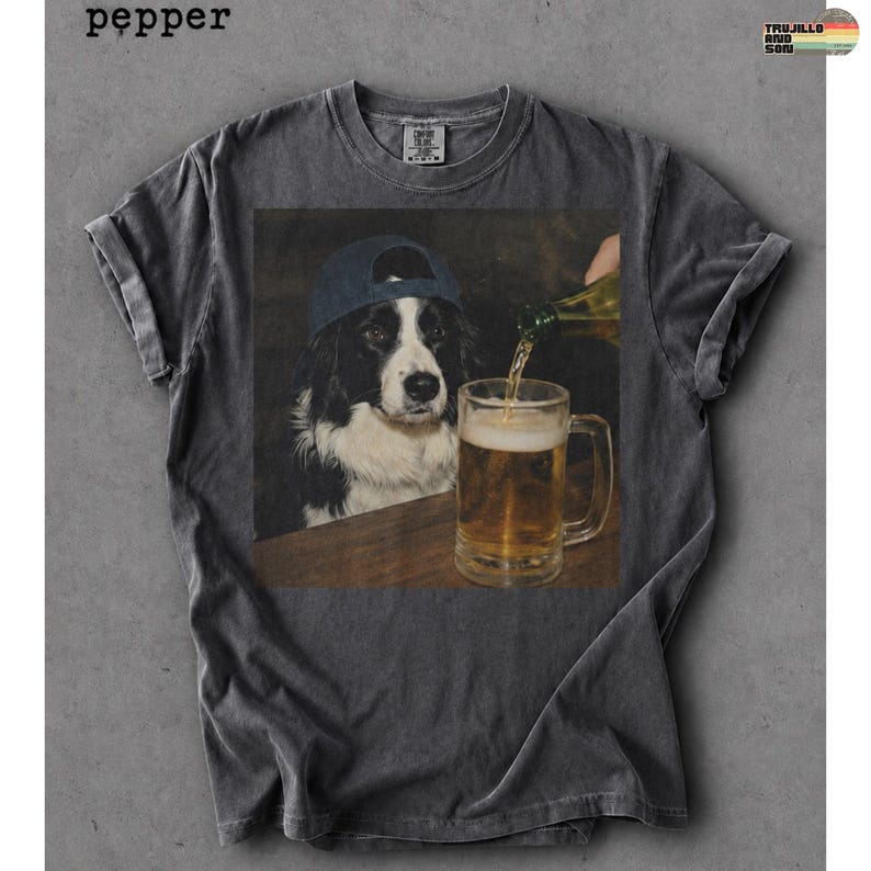 May include: A charcoal gray short-sleeve t-shirt featuring a graphic print of a black and white dog wearing a blue baseball cap, watching as liquid is poured from a bottle into a glass beer mug. The word pepper is printed in the top left corner.