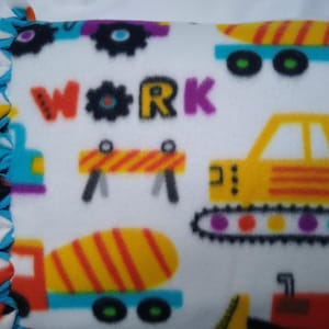 May include: A white fleece blanket with a colorful construction theme. The blanket features various construction vehicles, including a dump truck, a cement mixer, and a bulldozer. The word "WORK" is printed in colorful letters on the blanket.