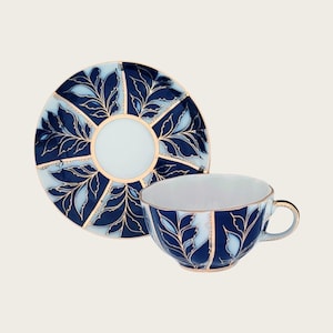 May include: A blue and white teacup and saucer set with gold leaf accents. The design features stylized leaves and a geometric pattern.