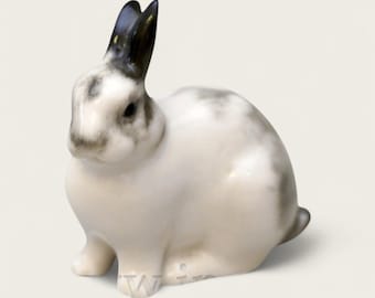 May include: A ceramic rabbit figurine in a seated position. The rabbit is primarily white with gray and black markings on its ears and back. The figurine has a glossy finish and detailed features, showcasing a realistic depiction of a rabbit.