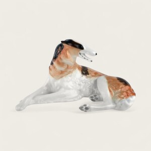 Może przedstawiać: Ceramiczna figurka psa Borzoi w pozycji spoczynku. Pies ma białe ciało z brązowymi, czarnymi i płowymi plamami sierści. Figurka ma długi, smukły pysk i leży.