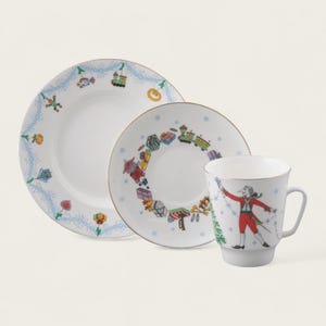 Puede incluir: Un juego de taza y platillo de porcelana blanca con temática navideña de invierno. El platillo presenta un diseño circular de regalos, un tren y un árbol de Navidad. La taza presenta a un hombre con un abrigo rojo sosteniendo una cadena de luces.