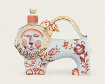 May include: A light blue ceramic lion-shaped teapot with gold accents and floral patterns. The lion has a detailed face with a red mane and a decorative top. The handle and feet are also adorned with gold.