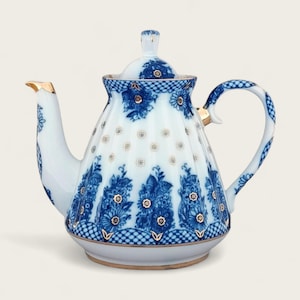 Puede incluir: Una tetera de porcelana blanca con un patrón floral azul y dorado. La tetera tiene un cuerpo redondo con un pico acampanado y un asa.