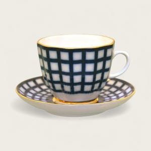 May include: A white teacup and saucer set with a navy blue grid pattern. The cup has a gold rim and a curved handle. The saucer also has a gold rim. The set is on a white background.