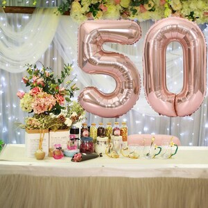 May include: Two large rose gold foil balloons in the shape of the numbers 50. The balloons are hanging from a white sheer curtain backdrop.
