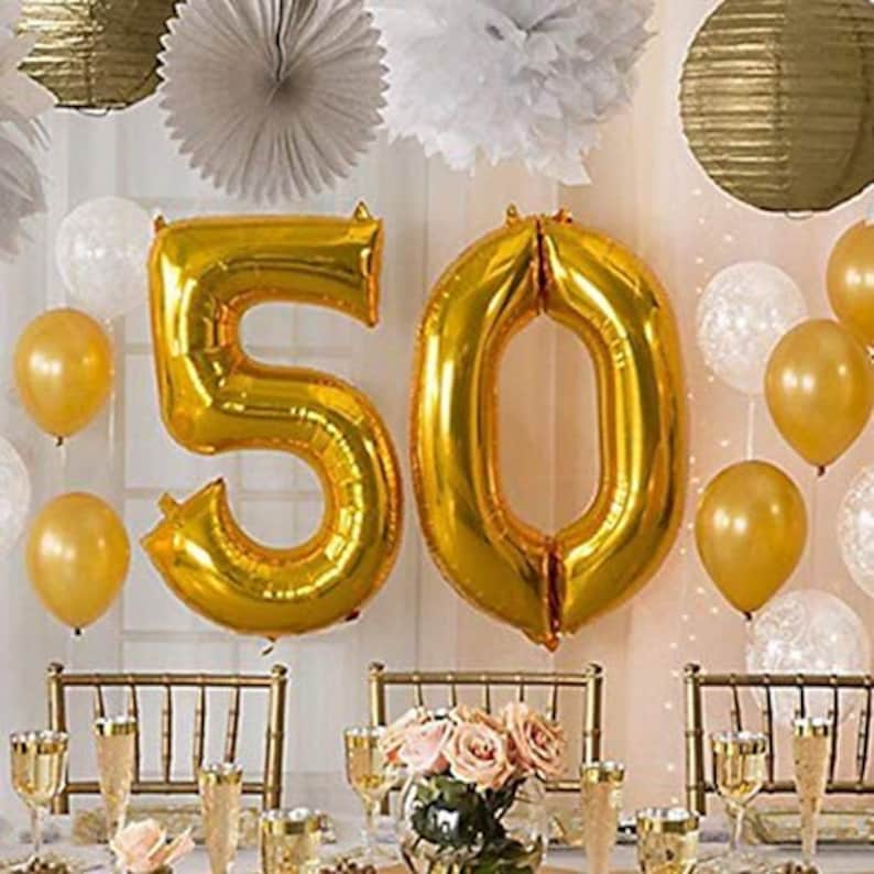 May include: Gold foil balloons spelling out the number "50" with gold and white balloons surrounding them. The balloons are in front of a white wall with gold and white decorations.