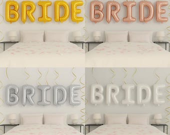 Könnte beinhalten: Vier Sätze von "BRIDE"-Buchstabenballons in Gold, Roségold, Silber und Weiß. Die Ballons hängen über Betten mit weißer Bettwäsche und kleinen Nachttischen. Der Hintergrund ist eine hellfarbige Wand mit goldenen Luftschlangen.