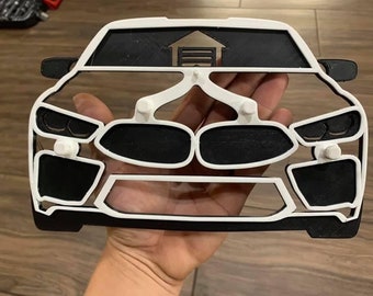 Op de afbeelding: Witte en zwarte omtrek van een auto met een garagedeur bovenop. De auto is van voren te zien en de garagedeur is gesloten. De omtrek is van plastic en dient als wanddecoratie.