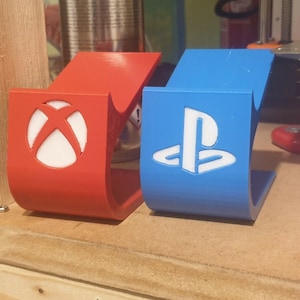 Puede incluir: Dos soportes para mandos impresos en 3D, uno rojo con el logotipo de Xbox y otro azul con el logotipo de Playstation.