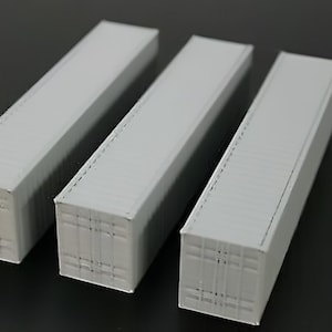 May include: Three gray miniature shipping containers arranged on a dark surface. The containers are rectangular with ridged tops and detailed sides, suggesting a model or toy.