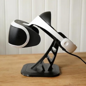 Puede incluir: Un soporte de auriculares VR negro con unos auriculares blancos apoyados en él. El soporte tiene un diseño geométrico y está hecho de plástico. Los auriculares tienen un diseño blanco y negro y están conectados a un cable negro.