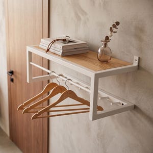 Pode incluir: Uma prateleira de parede em metal branco com uma superfície superior em madeira clara e um varão inferior. Três cabides de madeira estão pendurados no varão. A prateleira contém uma pilha de livros e um pequeno vaso de vidro.