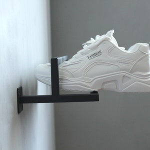 May include: A white sneaker with black accents and the word "FASHION" printed on the side is displayed on a black metal shoe rack mounted to a white wall.