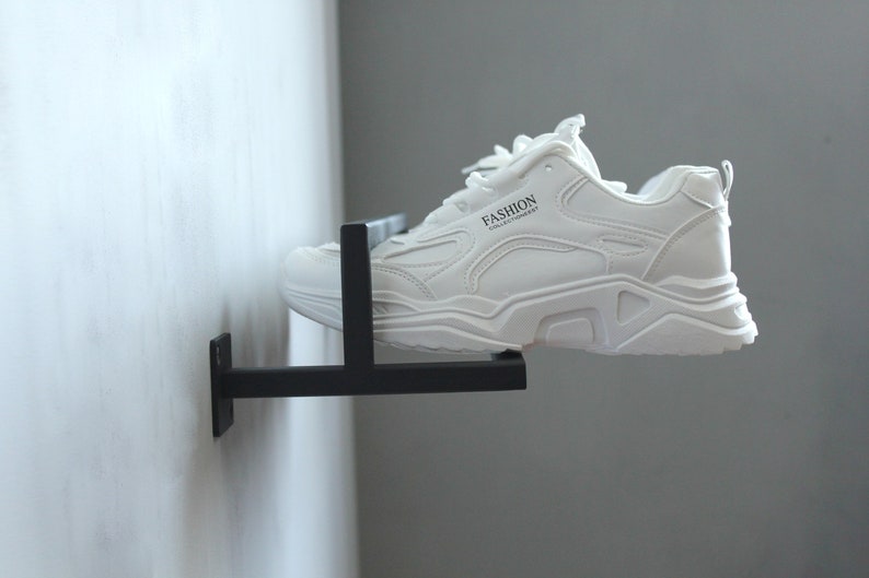 May include: A white sneaker with black accents and the word "FASHION" printed on the side is displayed on a black metal shoe rack mounted to a white wall.