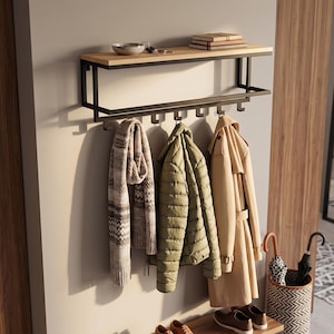 May include: A wall-mounted coat rack featuring a rectangular black metal frame with a light wood top shelf. The unit includes seven black metal hooks underneath for hanging items. A patterned scarf, a quilted olive jacket, and a tan trench coat hang.