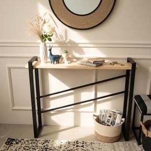 May include: A console table with a light wood top and a black metal frame featuring two diagonal support bars. The table holds a white vase with dried flowers, a blue deer figurine, a small succulent, two notebooks, and a set of metal keys.