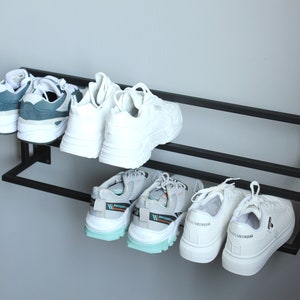 May include: A black metal shoe rack with two shelves mounted on a white wall. The rack holds six pairs of sneakers, including white, blue, and gray sneakers.