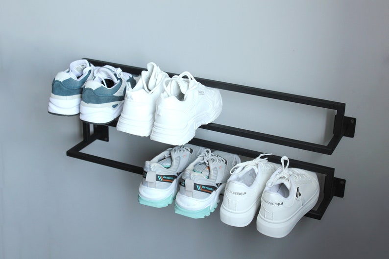 May include: A black metal shoe rack with two shelves mounted on a white wall. The rack holds six pairs of sneakers, including white, blue, and gray sneakers.