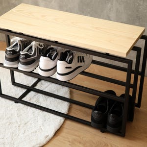以下が含まれることがあります： 3 段の棚と木製天板が付いた黒い金属製の靴棚。靴棚にはスニーカーやサンダルなど、さまざまな種類の靴が置かれています。