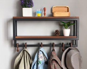 May include: A black metal wall-mounted shelf with a wooden top and hooks. The shelf has a small wooden box, a glass with a blue and yellow liquid, and a potted plant on the top shelf. There are three jackets and a hat hanging on the hooks.